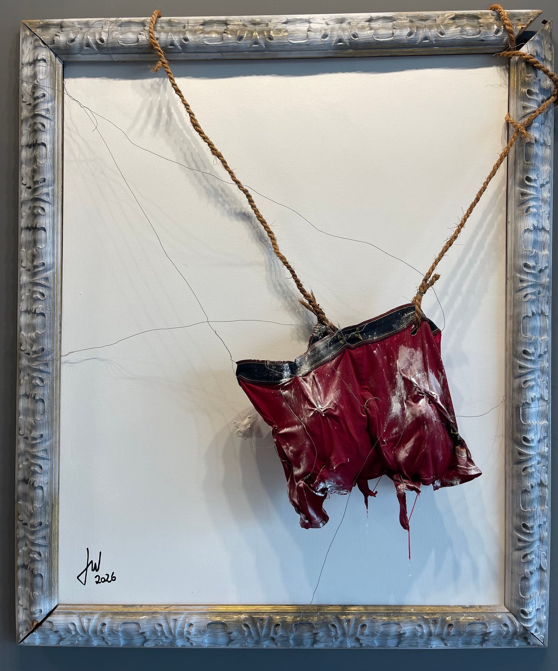 A deep red fabric hanging from a twisted rope inside an ornate silver-gray frame mounted on white wall background
