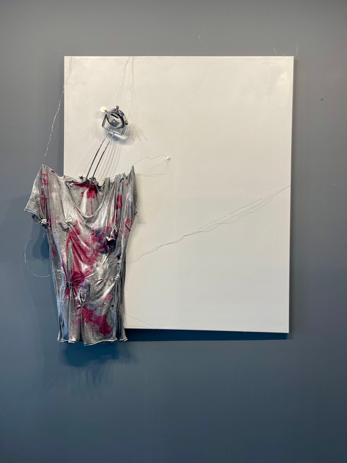 Plastic-wrapped artwork with pink and maroon patterned fabric suspended in front of a blank white canvas on a gray wall